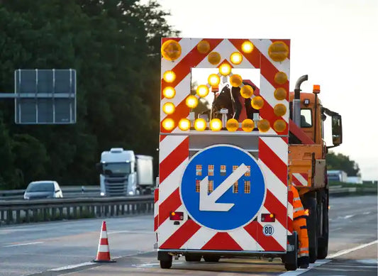 Absicherungsfahrzeug mit Warnpfeil und Rundumleuchten auf der Autobahn während einer Baustellenabsicherung; daneben steht eine Person in orangefarbener Warnkleidung, Verkehr läuft weiter im Hintergrund.
